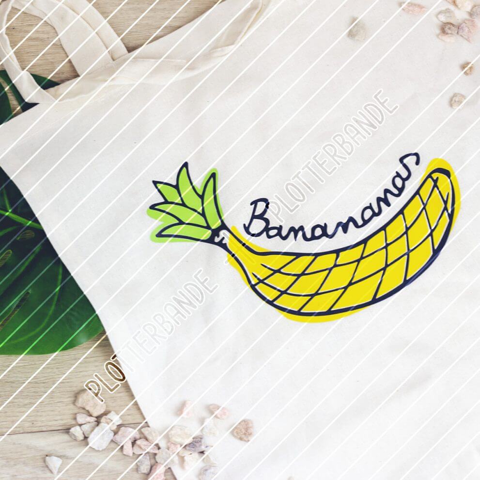 Auf einem Holzboden liegt ein weißer Beutel mit dem Banananas-Design der Plotterbande – eine Illustration, die Elemente einer Banane und einer Ananas verbindet.