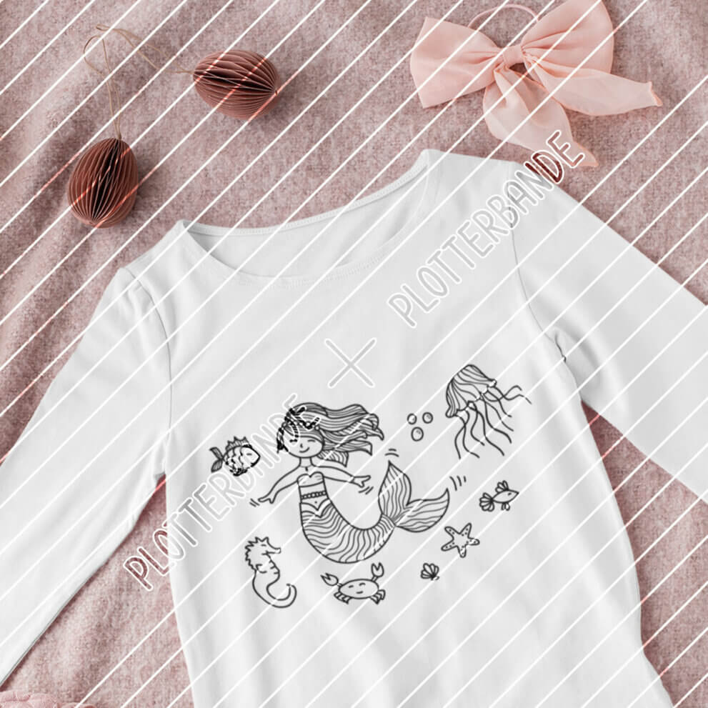 (1) Man sieht ein weißes T-Shirt auf einer rosa Decke. Auf dem Shirt ist das Meerjungfrau-Design der Plotterbande abgebildet.